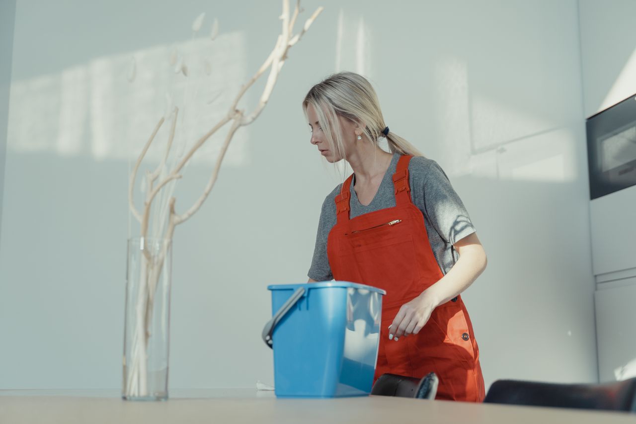 Une femme en uniforme rouge nettoie à l’intérieur avec un seau bleu, en veillant à maintenir un environnement impeccable.