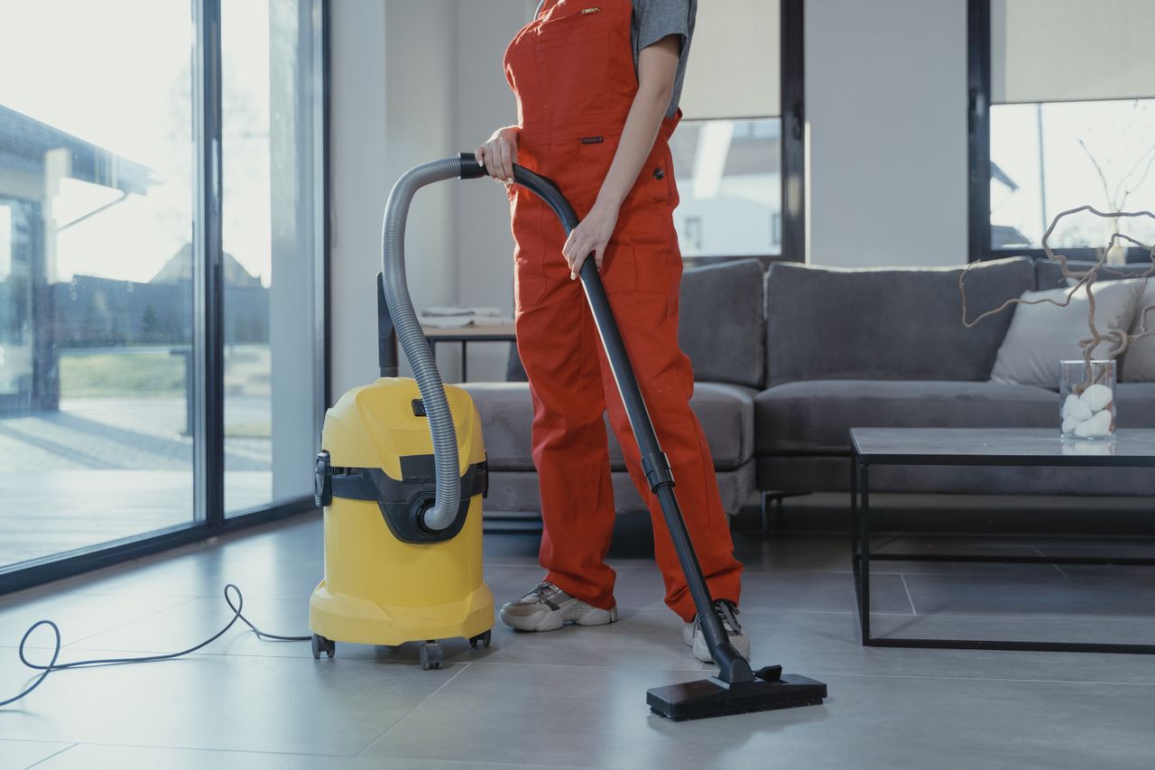 Un professionnel du nettoyage en salopette rouge aspirant un salon moderne avec un aspirateur jaune.